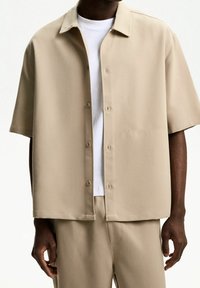 Beżowa koszula krótki rękaw z guzikami, z kołnierzykiem, kieszenią z przodu i gładką fakturą, nałożona na białą koszulkę z okrągłym dekoltem.