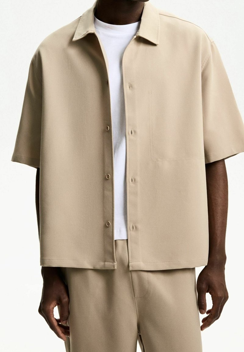 Beżowa koszula krótki rękaw z guzikami, z kołnierzykiem, kieszenią z przodu i gładką fakturą, nałożona na białą koszulkę z okrągłym dekoltem.