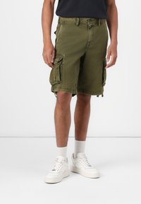Olive zöld cargo short, több oldalsó zsebbel, laza szabással és rojtos szegéllyel. Fehér sportcipővel és térdzoknival párosítva.