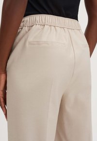 Beige broek met een elastieken tailleband, twee achterzakken en een gladde textuur. Het ontwerp heeft een ontspannen pasvorm, geschikt voor casual kleding.