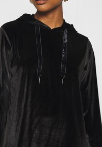 Felpa con cappuccio in velluto nero con lacci in satin. Presenta una texture morbida e una vestibilità rilassata, adatta per l'abbigliamento casual.