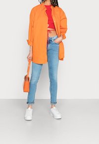 Persona con una camisa de gran tamaño de color naranja brillante, un top corto rojo, jeans ajustados de color azul claro, zapatillas de plataforma blancas y sosteniendo un bolso naranja.