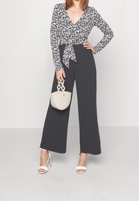 Top cruzado de patrón en blanco y negro con mangas largas, combinado con pantalones de pierna ancha en gris oscuro. Accesorios incluyen un bolso ligero en color beige y tacones blancos.