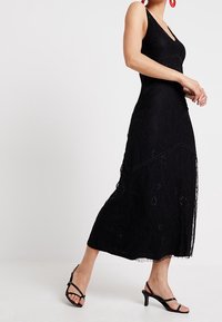 Černé krajkové maxi šaty s upnutým korzetem a rozšířenou sukní. Mají tenké ramínka, květinové vzory a vrstvenou texturu. Nosené s černými sandály.