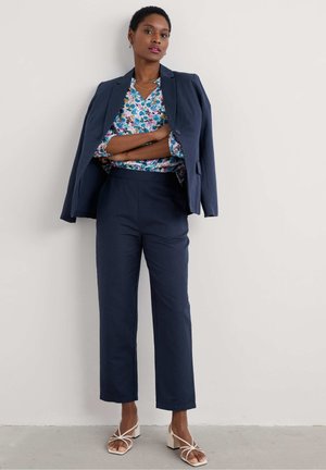 Marineblazer über einer Blumenshirt, kombiniert mit marineblauen Hosen und weißen Blockabsatzsandalen. Glatte Stoffe, maßgeschneiderte Passform und Farbakzente.