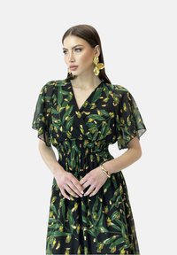 Schwarzes Kleid mit grünem und gelbem Blumenmuster, kurzen Schmetterlingsärmeln, elastischem Taillenbund, fließendem Rock, accessoriert mit großen goldenen Ohrringen.