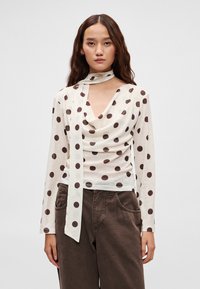 Kremowa bluzka z długim rękawem w brązowe groszki, z drapowanym dekoltem i dopasowanym szalikiem jako detalem. Miękka, lekka tkanina o luźnym kroju.