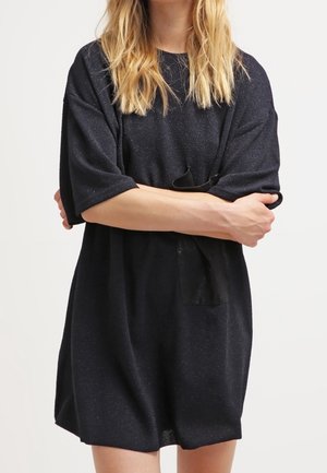 Femme aux longs cheveux blonds portant une robe noire ample à manches courtes, les bras croisés sur la poitrine, debout devant un fond blanc.