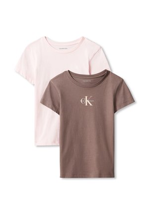 Ung pige med langt hår iført en brun Calvin Klein t-shirt, lyseblå jeans og sorte sko, stående mod en ensfarvet baggrund.