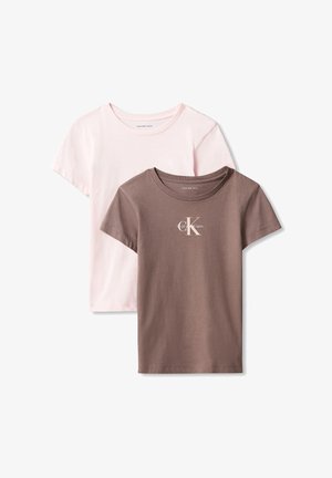 Młoda dziewczyna o długich włosach, ubrana w brązowy t-shirt Calvin Klein, jasnoniebieskie dżinsy i czarne buty, stoi na tle jednolitego tła.