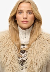 Un pull tricoté avec un col montant, orné d'un motif multicolore, superposé sous un gilet beige doux et duveteux.