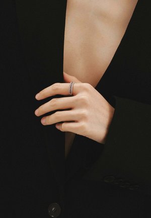 Anillo de plata con múltiples pequeñas piedras preciosas, banda lisa, sostenido por una mano, con una prenda negra de escote en V pronunciado de fondo.