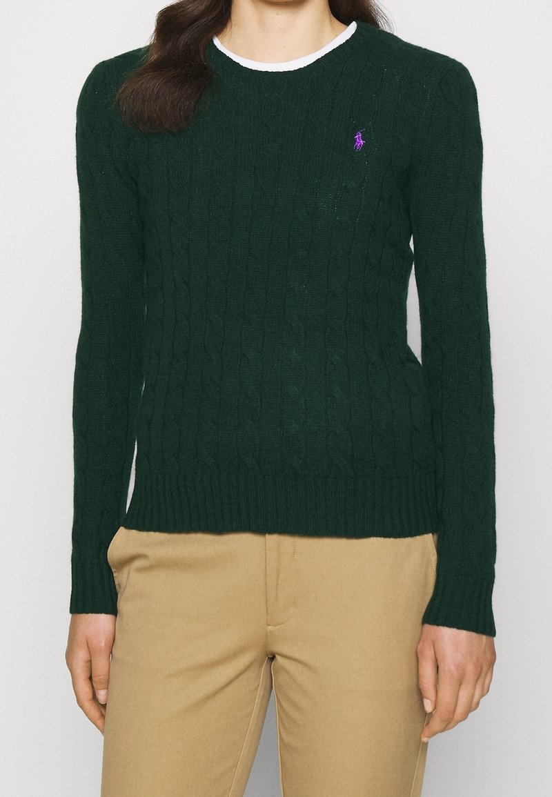 Femme portant un pull en maille torsadée vert foncé avec un logo violet sur le côté gauche de la poitrine, un pantalon beige et un sous-pull blanc visible au niveau du col.