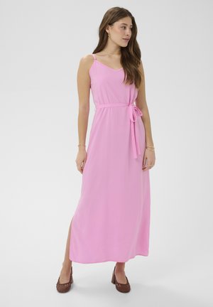 Kvinde iført ærmeløs lyserød maxikjole med sideslids og bundet taljebælte, parret med brune hælede sko, står på neutral baggrund.