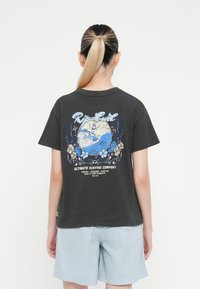 Črna bombažna majica s pisanimi grafičnimi valovi, logotipom "Rip Curl" in cvetličnimi detajli na hrbtni strani. V kombinaciji z svetlo modrimi patterned kratkimi hlačami.