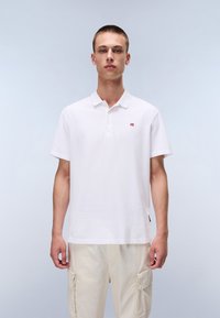 Fehér pamut póló rövid ujjakkal és háromgombos pamut nyakkivágással, a mellkason egy kis zászló emblémával.