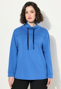 Felpa blu con colletto alto e cordini neri. Presenta maniche lunghe e una cucitura orizzontale testurizzata lungo il petto.