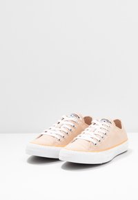 Ljust persikofärgade canvasgympaskor med vita snören, texturerad gummisula och orange accent. Klassisk rund tåkonstruktion, minimal branding på tungan.