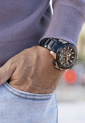 Montre noire et or rose avec un cadran rond, fonctions chronographe et un bracelet à maillons, portée sur un poignet avec un pull violet clair.