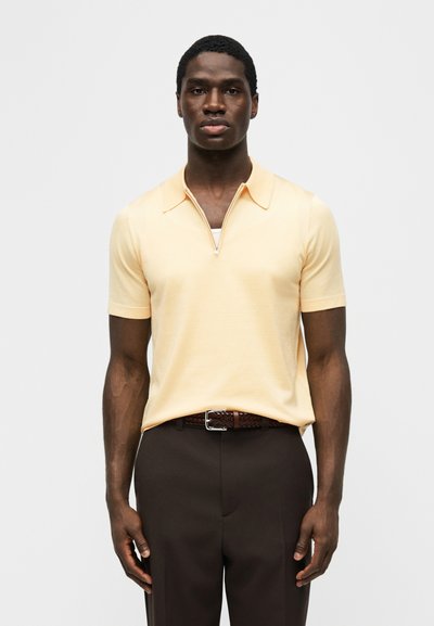 Άνδρας ντυμένος με απαλό κίτρινο κοντομάνικο πουκάμισο με φερμουάρ στον γιακά, που είναι μπουκλωμένο μέσα σε σκούρο καφέ παντελόνι με πλεγμένη καφέ ζώνη, στέκεται μπροστά σε λευκό φόντο.