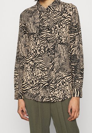 Personne portant une chemise boutonnée à manches longues avec un imprimé léopard et zèbre, associée à un pantalon vert olive, debout devant un fond uni.