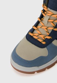 Nahaufnahme eines beigen, blauen und marineblauen Sneakers mit orangefarbenen und schwarzen gemusterten Schnürsenkeln auf einem weißen Hintergrund.
