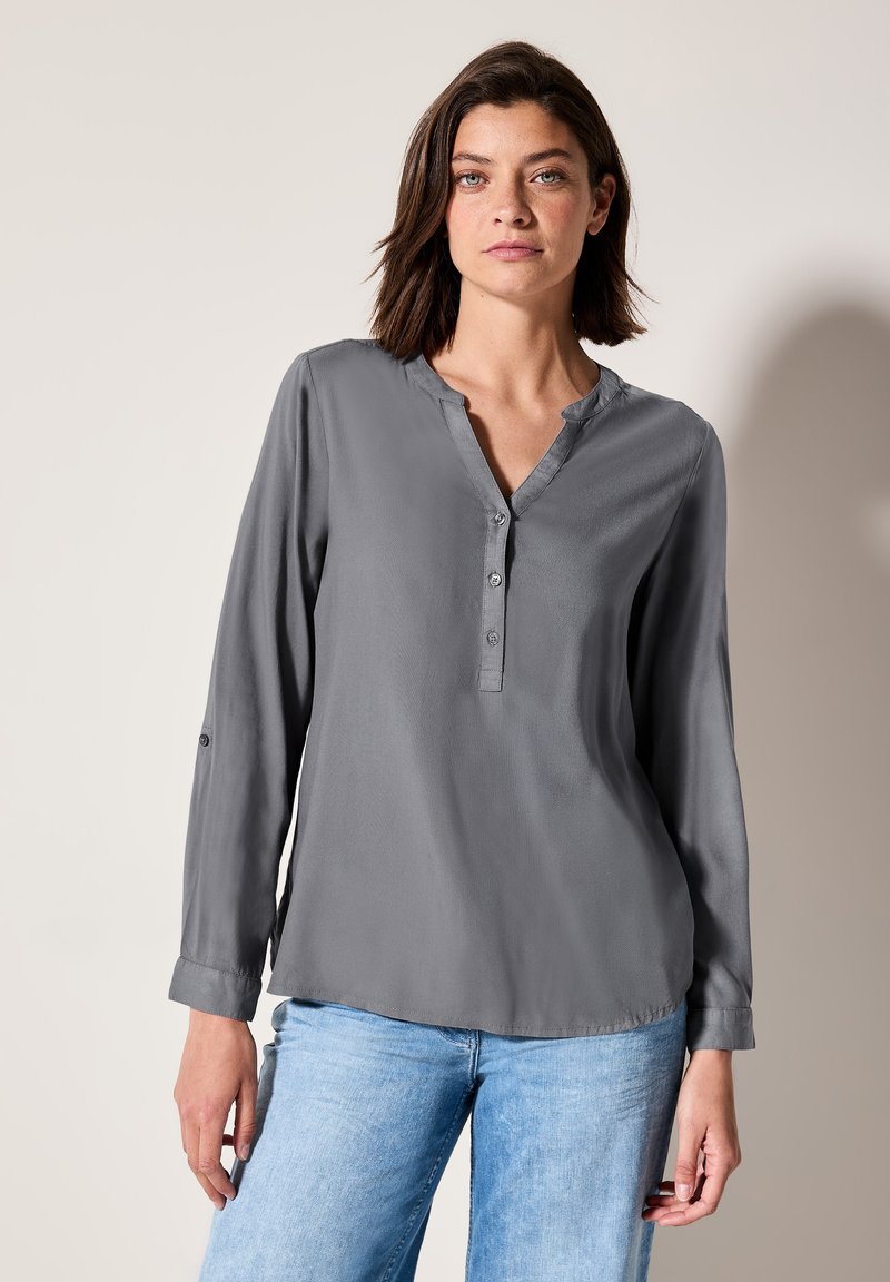 Blouse grise à manches longues avec un décolleté en V, patte de boutonnage à quatre boutons, texture douce et poignets à rouleau. Assortie avec un jean en denim bleu clair.