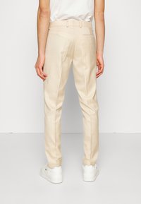 Pantaloni bej, croiți pe corp, cu o singură buzunar posterior și bucle pentru curea, asortați cu adidași albi pe un fundal uniform.