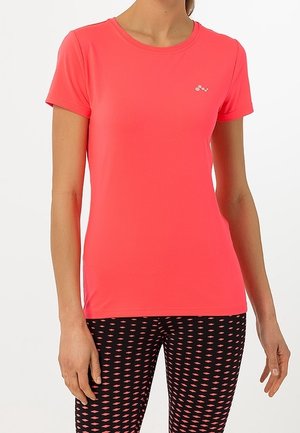 Kobieta ubrana w jasnoróżową, koralową sportową koszulkę z krótkim rękawem oraz czarne legginsy z koralowym wzorem w romby, stojąca na białym tle.