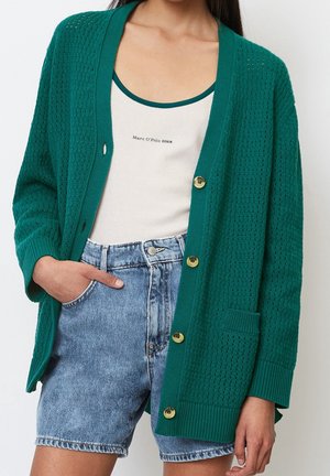 Vrouw draagt een groene, gestructureerde cardigan met grote knopen over een wit tanktopje en een highwaisted blauwe denim korte broek, met één hand in de zak.