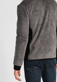 Person klädd i en grå fleecjacka med svarta sidopaneler och mörkblå jeans, sedd bakifrån mot en enfärgad bakgrund.