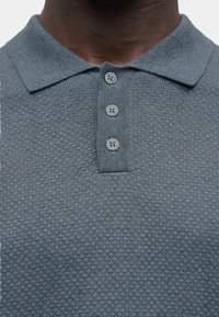 Nahaufnahme einer dunkelhäutigen Person, die ein blaues, strukturiertes Poloshirt mit einer dreiknöpfigen Knopfleiste und einem weichen Kragen trägt.