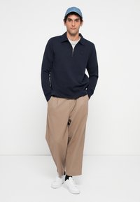 Man wearing navy zip-up long sleeve shirt, beige wide-leg pants, white sneakers, and blue cap standing with hands in pockets.