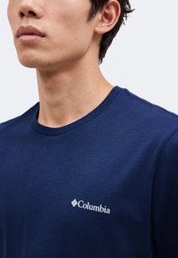 Άτομο που φοράει μπλε σκούρο T-shirt Columbia, δείχνοντας το λογότυπο στο αριστερό στήθος με κοντινό πλάνο στο λαιμό και το σαγόνι.