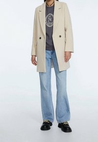 Persona vestida con un abrigo beige de doble botonadura, sudadera gráfica azul marino, vaqueros de pierna ancha de color azul claro y zapatos negros con cadenas doradas.