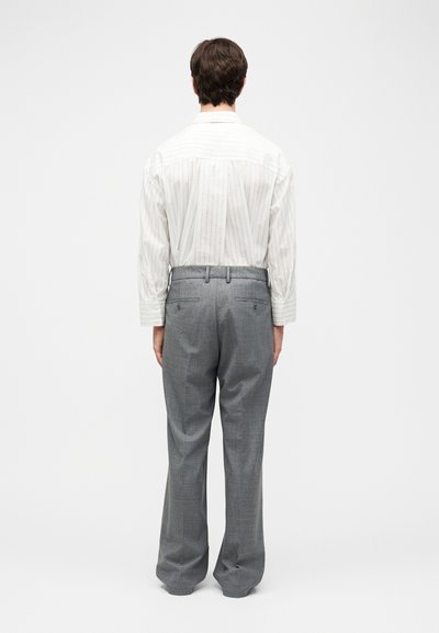 Άνδρας που στέκεται με την πλάτη, φοράει λευκό ριγέ πουκάμισο και γκρι ραμμένο στο μέτρο παντελόνι μπροστά σε απλό φόντο.