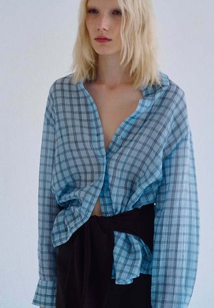 Mujer rubia con camisa de cuadros azul suelta con botones y cinturón de tela negro delante, de pie frente a un fondo claro y liso.