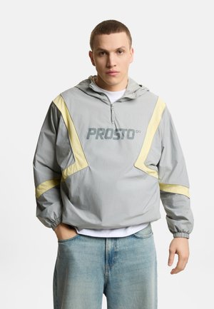 Młody mężczyzna w szaro-żółtej, z kapturem wiatrówce z logo „PROSTO”, jasnoniebieskich dżinsach, stoi na jednolitym białym tle.