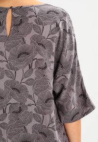 Blusa floreale grigia con vestibilità ampia, caratterizzata da pattern di fiori contornati in nero e un dettaglio a chiave sul retro. Maniche corte e larghe.