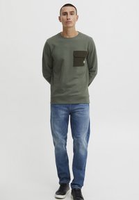 Green crewneck sweatshirt with a chest pocket, paired with blue denim jeans and black sneakers. The sweatshirt features a relaxed fit.