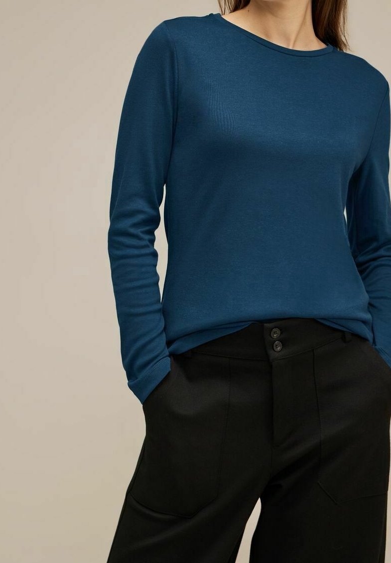 Chemise en teal à manches longues en tissu doux, avec un col rond, associée à un pantalon noir ajusté avec une fermeture à bouton et des poches.