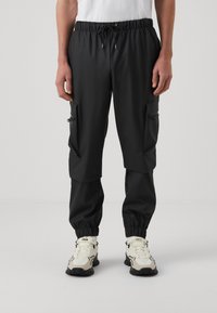 Pantaloni cargo neri con polsini elasticizzati, vita regolabile e molteplici tasche laterali con zip. Il materiale sembra leggero e liscio.
