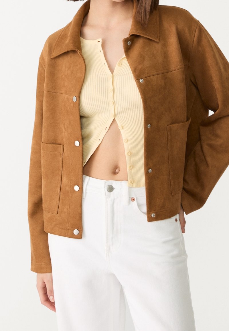 Brun jakke i imiteret ruskind med krave, trykknapper og to forlommer, kombineret med en cremefarvet ribbet top og hvide jeans.