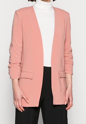 Person trägt ein hellrosa Sakko über einem weißen, gerippten Rollkragenpullover und schwarze Hosen, steht vor einem schlichten hellen Hintergrund.