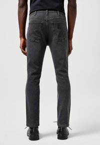 Moški, ki nosi zbledene črne Wrangler jeans in črne čevlje, prikazan od zadaj na enostavni svetli podlagi.