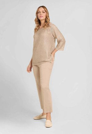 Donna con capelli mossi, in piedi, indossa un maglione beige a maglia, pantaloni beige e scarpe slip-on beige chiaro su uno sfondo chiaro e uniforme.