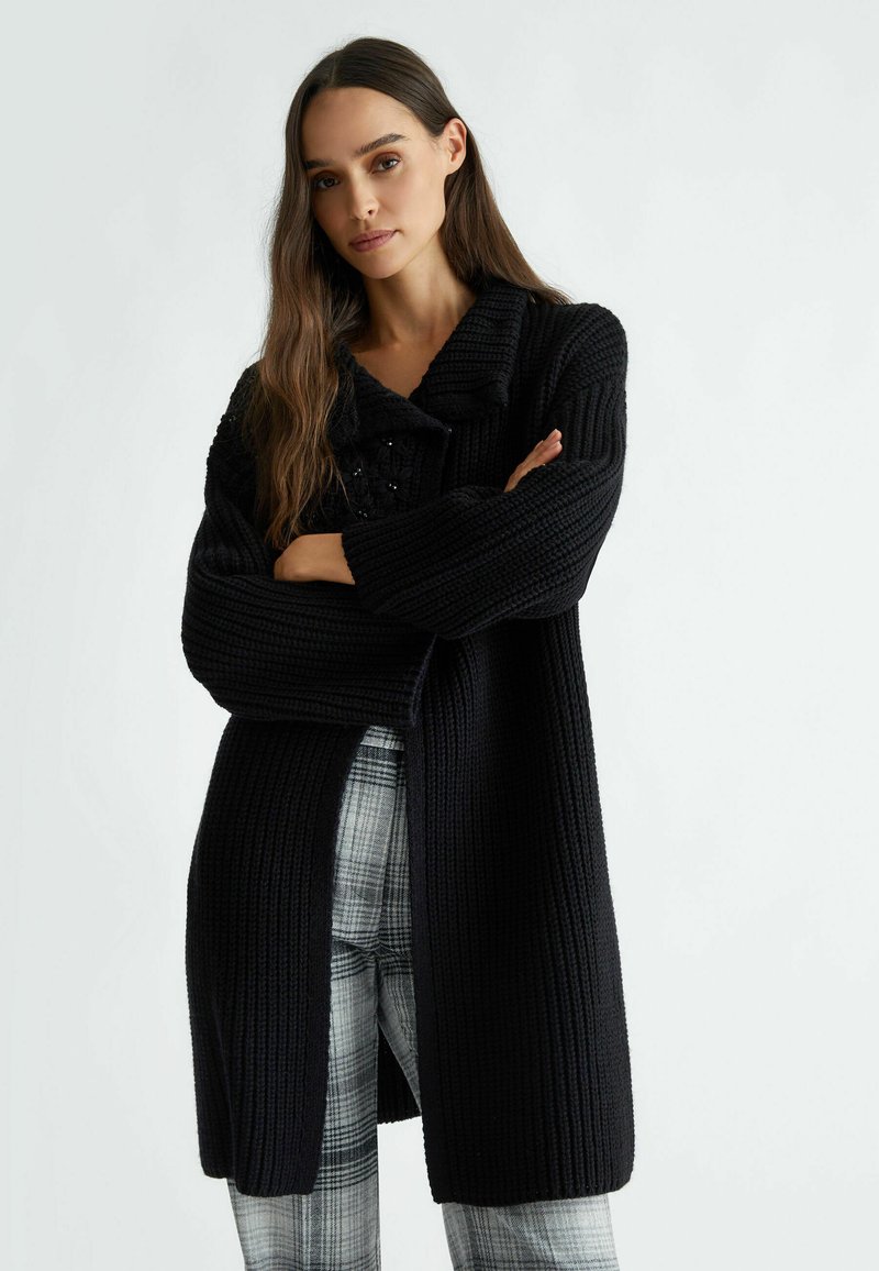 Černý pletený kardigan s výrazným límcem, dlouhými rukávy, žebrovanou texturou a otevřeným předním dílem, doplněný černobílými kostkovanými kalhotami.