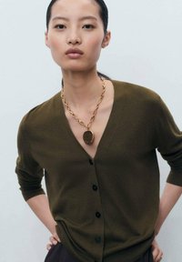Olive green cardigan with a deep V-neck, long sleeves, and black buttons. Accented by a gold chain necklace with a round pendant.