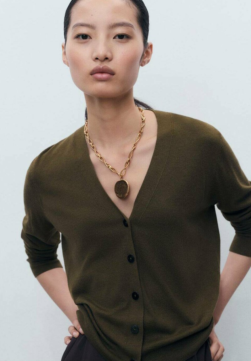 Olive green cardigan with a deep V-neck, long sleeves, and black buttons. Accented by a gold chain necklace with a round pendant.