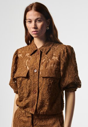 Junge Frau mit langen roten Haaren, die ein braunes Spitzenoberteil mit kurzen Puffärmeln und passende Unterteile trägt, blickt nach vorne vor einem einfarbigen Hintergrund.